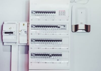 Gros plan sur un tableau électrique moderne et conforme fixé sur un mur blanc. Le tableau est composé de plusieurs rangées de disjoncteurs et de modules de protection, ainsi que d'un compteur électrique et d'un disjoncteur général sur la gauche. Sur la droite, deux appareils de domotique ou d'alarme sans fil sont installés.