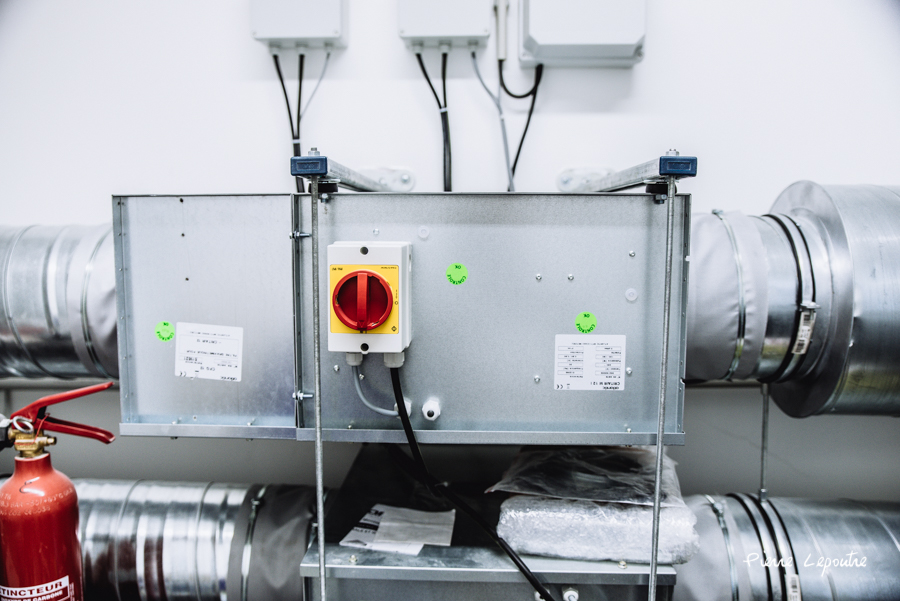 Caisson de traitement d’air (CTA) et alimentation électrique d’une installation de ventilation. Gros plan sur un caisson métallique de traitement d'air ou de ventilation, monté sur un mur blanc. Un interrupteur principal de couleur rouge et jaune y est fixé.