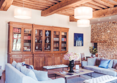 Salon élégant d'un hôtel, mêlant rustique et moderne. Le plafond est caractérisé par de grandes poutres en bois apparentes. Des canapés clairs, une bibliothèque ancienne en bois remplie de livres et de décorations, et un mur en pierres apparentes créent une ambiance chaleureuse. De grands luminaires cylindriques diffusent une lumière douce.