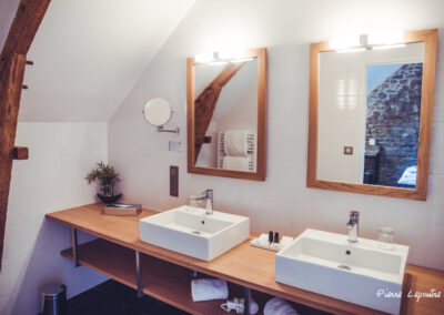 Salle de bain élégante avec double vasque. Deux lavabos rectangulaires blancs sont posés sur un long plan de travail en bois clair. Au-dessus, deux miroirs encadrés de bois avec éclairage intégré reflètent une poutre apparente et un mur en pierre. Le mur est carrelé en blanc et le sol est sombre.