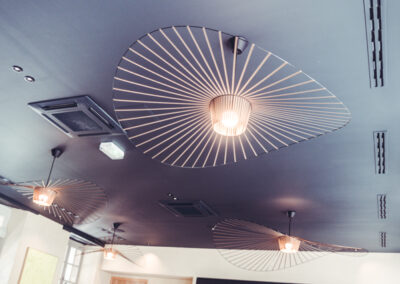 Vue d'angle plongeante sur l'intérieur élégant d'un restaurant d'hôtel 4 étoiles à Saint-Malo. Le plafond sombre est orné de luminaires design en forme d'éventail et intègre discrètement des bouches de climatisation.