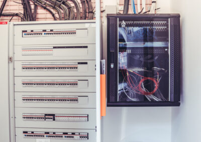 Tableau électrique et baie de brassage dans un local technique. À gauche, un grand tableau de distribution électrique modulaire gris est visible, avec ses disjoncteurs. À droite, une baie réseau noire fermée, contenant des câbles réseau. Des faisceaux de câbles électriques et de communication montent vers le plafond.