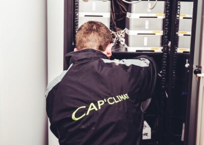 Vue de dos d'un technicien portant une veste noire avec le logo CAP'CLIMAT inscrit en vert. Il est penché sur une armoire de serveur ou un coffret de climatisation technique, où des modules (boîtiers gris) et un enchevêtrement de câbles sont visibles. Le technicien est en pleine intervention de maintenance ou d'installation.