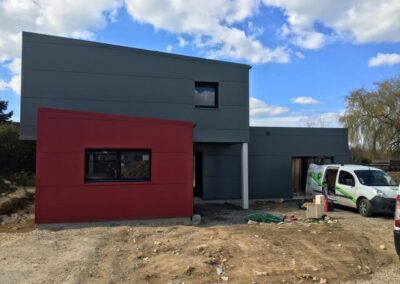 Vue extérieure d'un chantier de construction ou de rénovation, montrant un bâtiment moderne à toit plat avec un bardage de couleur gris anthracite et une extension ou un module d'entrée de couleur rouge vif. Le bâtiment présente une architecture avec des volumes décalés. L'environnement est en terre battue. Une fourgonnette de chantier blanche Cap'Climat est garée devant l'entrée.