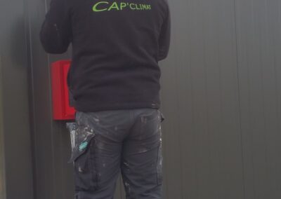 Un homme, dos à l'objectif, portant un pull polaire noir avec le logo vert "CAP CLIMAT", est debout sur un escabeau.