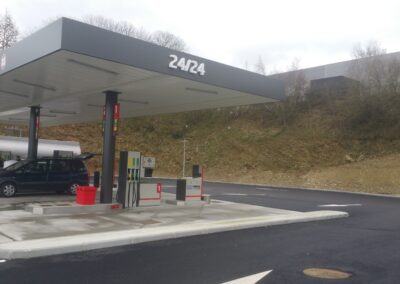 Une station-service moderne, sous un auvent gris et blanc affichant "24/24". Les pompes à essence sont installées sur une zone bétonnée et une voiture est en train de faire le plein.