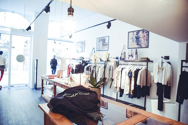 Vue intérieure d'une boutique de prêt-à-porter féminin au design élégant et lumineux. Les vêtements sont présentés sur des portants métalliques. Au centre, une grande table en bois de style ancien sert de présentoir. L'éclairage est assuré par un mélange de spots sur rail noir modernes, d'une grande ampoule suspendue de style industriel et d'une forte lumière naturelle provenant de la vitrine d'entrée. Une orchidée et des cadres photo décorent l'espace.