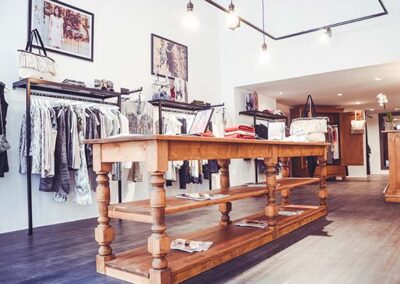 Vue intérieure d'une boutique de prêt-à-porter chic, avec un grand présentoir central en bois clair de style rustique/ancien. Autour de cette table, des portants noirs minimalistes exposent des vêtements. L'éclairage est assuré par des spots sur rail noir et des ampoules pendantes à filaments, créant une ambiance chaleureuse. Le sol est en parquet foncé et les murs sont blancs, décorés de cadres.