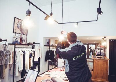 Vue de l'intérieur d'une boutique de prêt-à-porter où un technicien (portant une veste de travail avec le logo "CAP'CLIMAT") est en train de régler ou d'installer des suspensions lumineuses à grosses ampoules à filament au-dessus d'une table de présentation. L'éclairage est complété par des spots sur rails noirs.