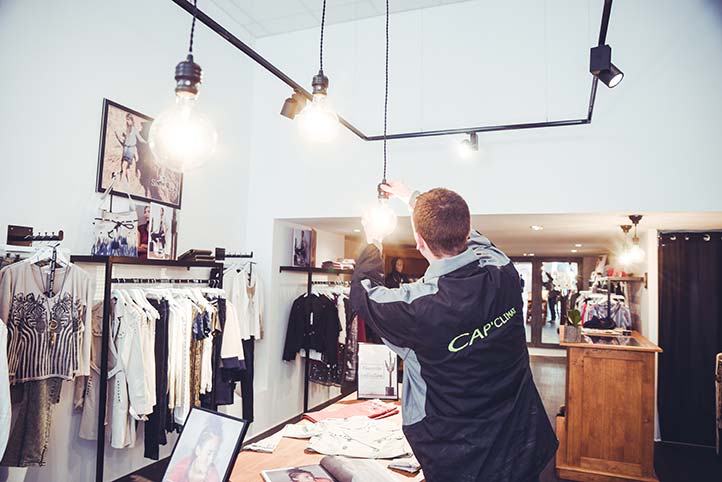 Vue de l'intérieur d'une boutique de prêt-à-porter où un technicien (portant une veste de travail avec le logo "CAP'CLIMAT") est en train de régler ou d'installer des suspensions lumineuses à grosses ampoules à filament au-dessus d'une table de présentation. L'éclairage est complété par des spots sur rails noirs.