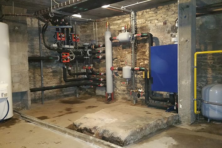 Rénovation technique : Le cœur de chauffe de l’hôtel de Nantes Vue intérieure d'une chaufferie rénovée dans un sous-sol aux murs en pierres apparentes et au sol en béton. L'installation est complexe et moderne, comprenant un réseau de tuyauteries isolées en noir et gris, des circulateurs rouges, des vannes, un ballon de stockage (blanc à gauche), et une chaudière/échangeur (caisson bleu à droite). L'ensemble des équipements est neuf et bien organisé.
