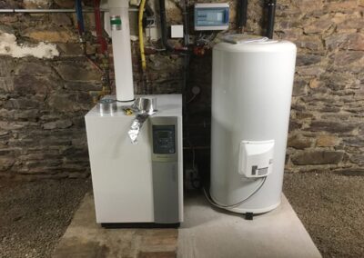 Gros plan sur une chaudière moderne et un ballon d'eau chaude (cumulus), tous deux de couleur blanche, installés sur une dalle de béton dans une pièce technique. Le mur derrière est un mur en pierres apparentes d'un sous-sol ou d'une ancienne bâtisse. Un tableau de commande numérique et une évacuation de fumée isolée sont visibles au-dessus des équipements.