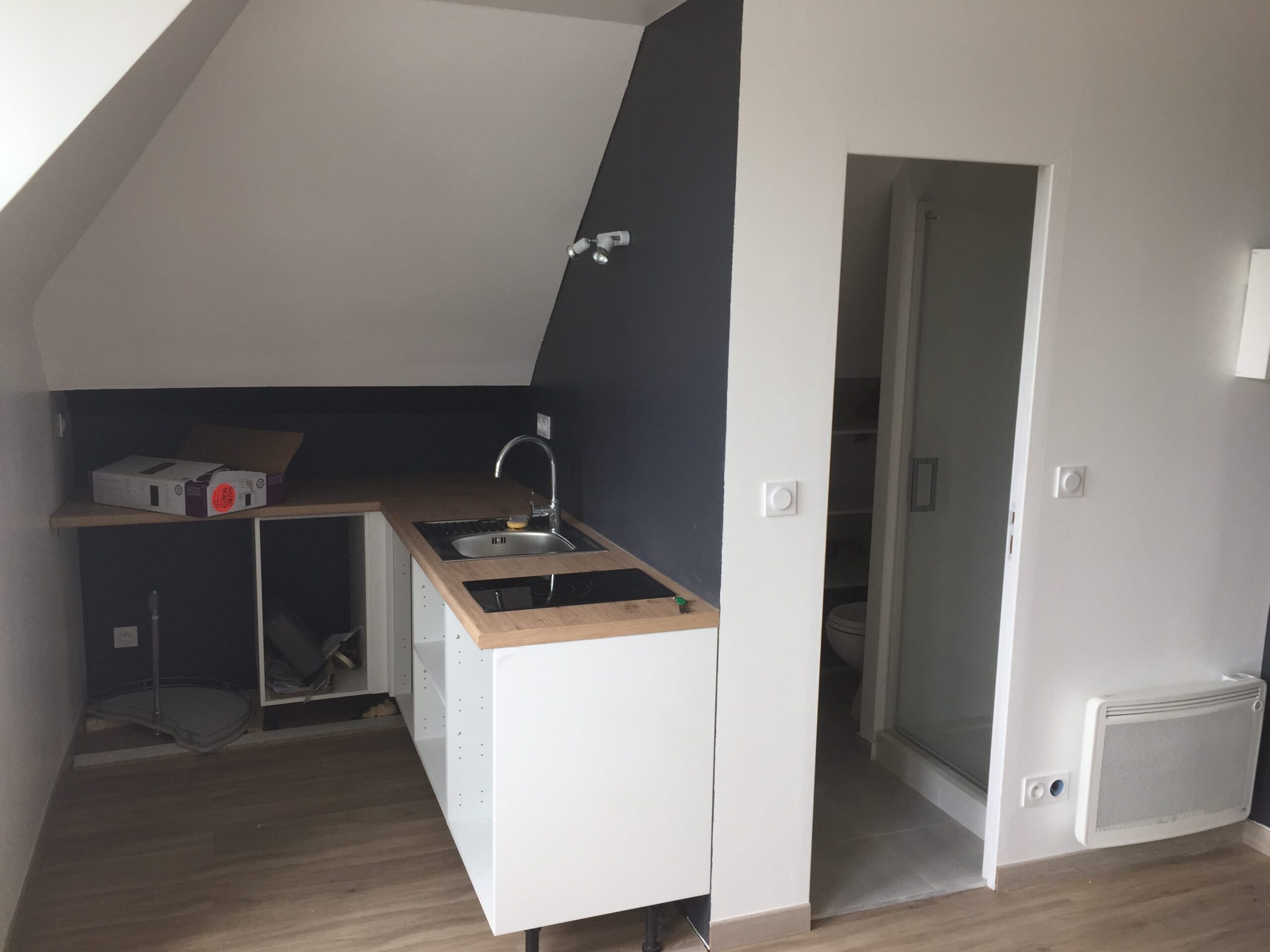 Vue intérieure d'un studio en cours de rénovation aménagé sous les combles. On y voit une kitchenette moderne en cours d'installation, avec des meubles bas blancs, un plan de travail en bois clair, un évier et des plaques de cuisson. Le mur derrière la kitchenette est peint en gris foncé (ou anthracite), contrastant avec les murs et les rampants blancs. À droite, l'entrée d'une salle d'eau avec une cabine de douche vitrée est visible.
