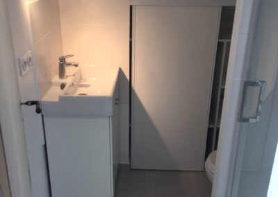 Vue intérieure d'une salle d'eau sous les combles à l'aménagement optimisé. Un meuble vasque blanc moderne est installé sous le rampant, avec un éclairage ciblé (spot mural). La pièce est carrelée de dalles grises. À droite de la vasque, un placard intégré avec porte coulissante dissimule l'espace WC et rangement, optimisant l'espace sous la pente du toit. L'entrée de la douche vitrée est visible à l'extrême droite.