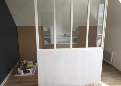 Vue intérieure d'un studio en cours de rénovation (sous les combles) montrant une cloison style verrière d'atelier peinte en blanc. Cette verrière sépare le séjour d'un espace de couchage ou de rangement. Un mur d'accent gris foncé est visible à gauche. L'espace est optimisé avec des placards bas en bois intégrés sous le rampant du toit.