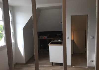 Vue intérieure d'un studio en cours de rénovation (sous les combles) prise à travers l'encadrement d'une porte. Au centre, on aperçoit une kitchenette minimaliste en cours d'installation, avec un évier et un plan de travail. La zone cuisine est sombre, sous le rampant du toit. L'espace est très lumineux grâce à une fenêtre sur la gauche. Le sol est en parquet stratifié clair. Un reflet de miroir accentue la perspective.