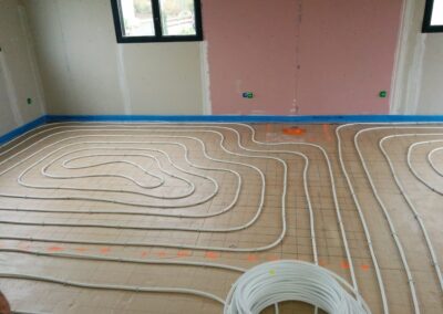 Vue intérieure d'une pièce en construction (murs en placo-plâtre) montrant l'installation d'un plancher chauffant hydronique. Un réseau de tubes blancs (circuits de chauffage) est soigneusement disposé en spirales sur un isolant au sol, fixé à l'aide d'un treillis. Un rouleau du tube non déroulé est visible au premier plan.