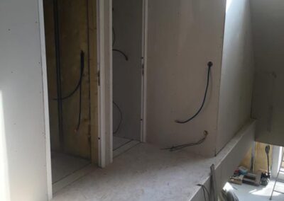 Vue intérieure d'un étage ou d'une mezzanine dans une maison en travaux (maison passive à Vitré). Le sol est recouvert de panneaux OSB. Les murs sont en plaques de plâtre (placo) non peintes, avec des gaines électriques en attente. Deux encadrements de portes sont visibles à gauche, et on aperçoit derrière l'un d'eux l'isolation en laine de roche ou de verre. La lumière naturelle est abondante, provenant de la hauteur. Un technicien est partiellement visible en bas à droite, travaillant sous la mezzanine.