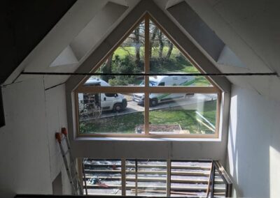 Vue intérieure d'une maison en construction, prise depuis un étage supérieur, montrant une grande fenêtre triangulaire et rectangulaire sous la charpente du toit. L'encadrement en bois met en valeur la vue sur la nature à l'extérieur. Le verre reflète deux fourgons de chantier garés. Les murs sont recouverts de plaques de plâtre (placo) non peintes. En bas, on voit la zone du futur plancher avec la structure en bois et un échafaudage.