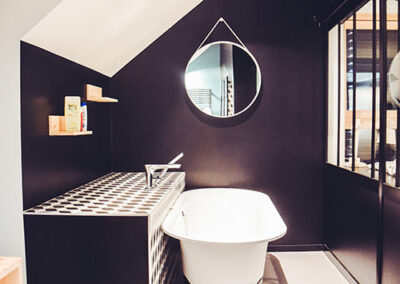 Vue d'une salle de bain moderne aménagée sous les combles, jouant sur un contraste marqué entre le blanc et le noir (ou anthracite). Une baignoire rétro sur pieds de couleur blanche est placée contre un mur sombre. À côté, un meuble de rangement est surmonté d'un carrelage à damier noir et blanc. Un miroir rond avec une fine sangle de suspension est accroché au mur sombre. Une verrière intérieure foncée sépare la salle de bain d'une autre pièce.
