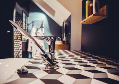 Gros plan sur une robinetterie moderne et chromée (douchette à main et mitigeur) installée sur le rebord d'une baignoire. Le rebord est recouvert d'un carrelage à motifs géométriques en relief noir, blanc et gris (effet 3D). L'arrière-plan flou montre l'intérieur d'une salle de bain sous les combles avec des murs sombres, une étagère en bois et un porte-serviettes chauffant.