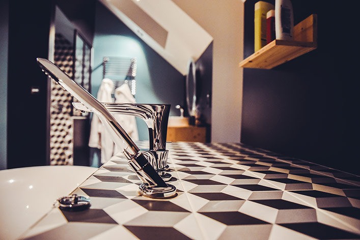 Gros plan sur une robinetterie moderne et chromée (douchette à main et mitigeur) installée sur le rebord d'une baignoire. Le rebord est recouvert d'un carrelage à motifs géométriques en relief noir, blanc et gris (effet 3D). L'arrière-plan flou montre l'intérieur d'une salle de bain sous les combles avec des murs sombres, une étagère en bois et un porte-serviettes chauffant.