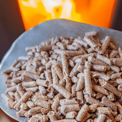 Une pelle métallique remplie de granulés de bois (pellets) tenue devant les flammes vives d'un poêle ou d'une chaudière à biomasse.