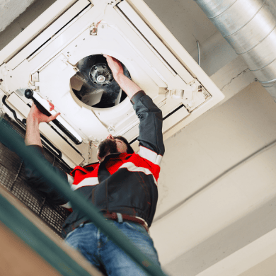 Un technicien travaille sur une unité de climatisation de type cassette encastrée au plafond, les mains à l'intérieur de l'appareil ouvert.