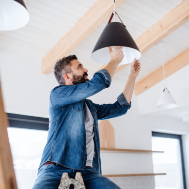 Un électricien professionnel, debout sur un escabeau, installe avec soin une ampoule dans une suspension moderne noire au plafond d'une maison neuve.