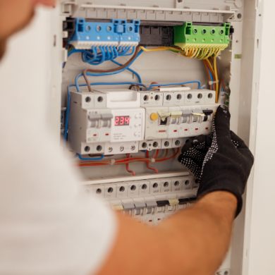 Un électricien portant un gant de protection noir intervient sur un tableau électrique moderne. On aperçoit un compteur numérique affichant 234V et plusieurs rangées de disjoncteurs parfaitement câblés avec des fils de différentes couleurs (bleu, vert, jaune, rouge).