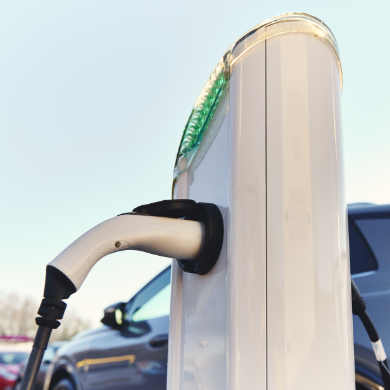 Vue en gros plan d'un pistolet de recharge blanc branché sur une borne de recharge électrique publique ou privée, avec une voiture garée à côté sous un ciel clair.