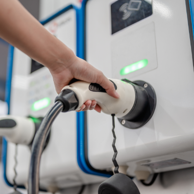ue rapprochée d'une main tenant un pistolet de recharge blanc et s'apprêtant à le brancher sur une borne de recharge murale. La borne présente un design moderne blanc et bleu avec des voyants LED verts allumés et un symbole de paiement sans contact.