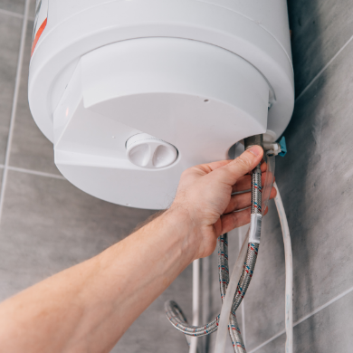 Vue rapprochée d'une main d'artisan vérifiant les raccordements hydrauliques flexibles sous un chauffe-eau électrique mural blanc. L'appareil est installé sur un mur avec un carrelage gris moderne.