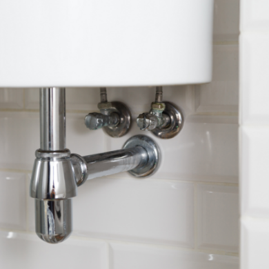 Vue en gros plan des raccordements chromés sous un lavabo blanc. On voit le siphon et les deux robinets d'arrêt fixés sur un mur en carrelage de type "métro" blanc. L'installation est propre, moderne et parfaitement alignée.