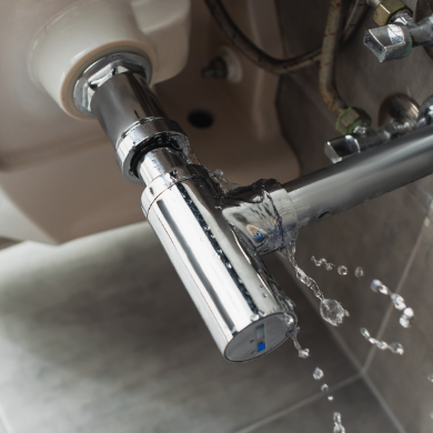 Fuite d'eau importante s'écoulant d'un siphon chromé sous un lavabo. Des gouttes d'eau sont projetées en l'air, illustrant un besoin urgent de réparation en plomberie.