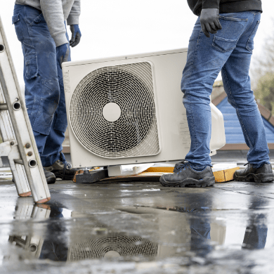 Deux techniciens portant des gants de protection déplacent avec précaution une unité extérieure de pompe à chaleur sur un toit plat mouillé. Une échelle est visible sur le côté.